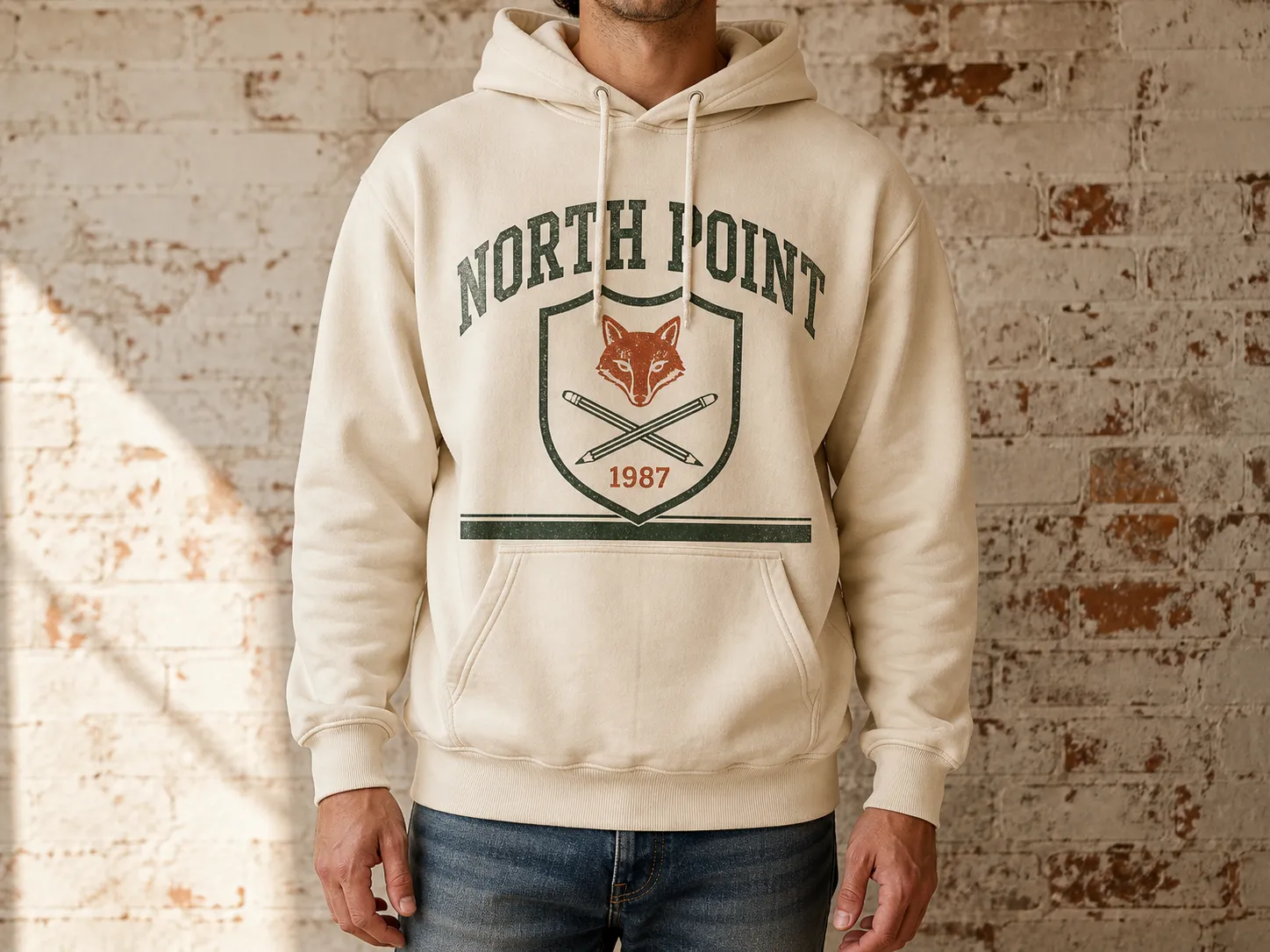 Man in a beige "North Point" hoodie with fox emblem and "1987" text, stands against a rustic brick wall, wearing jeans.