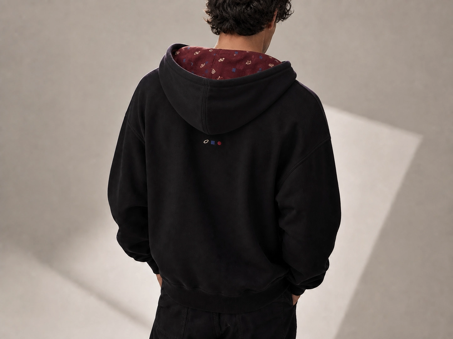 Man in a black hoodie with a patterned maroon lining, standing with hands in pockets, facing away in a softly lit room.