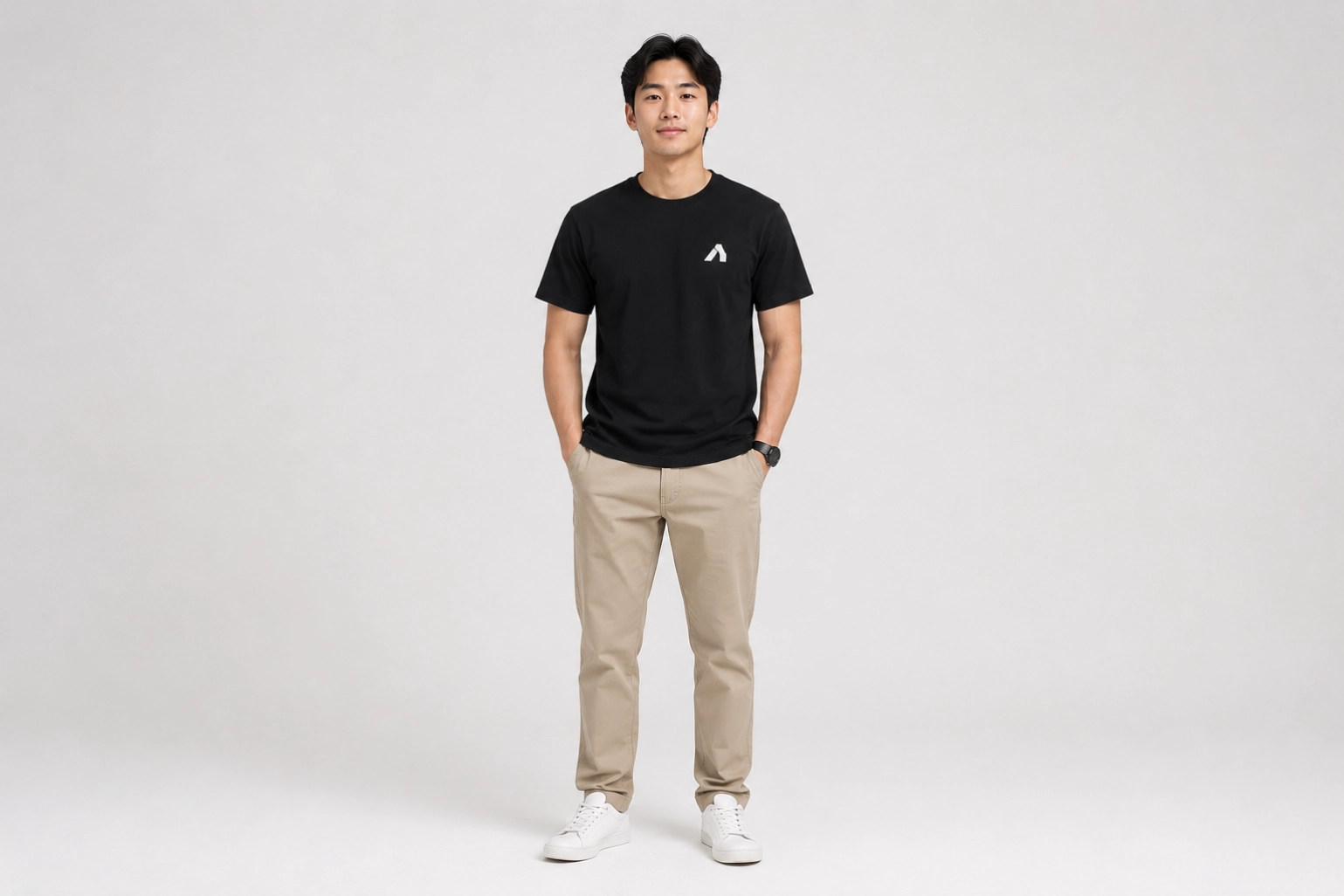 Man standing facing the camera in a black t-shirt with beige chinos and white sneakers against a light gray backdrop.