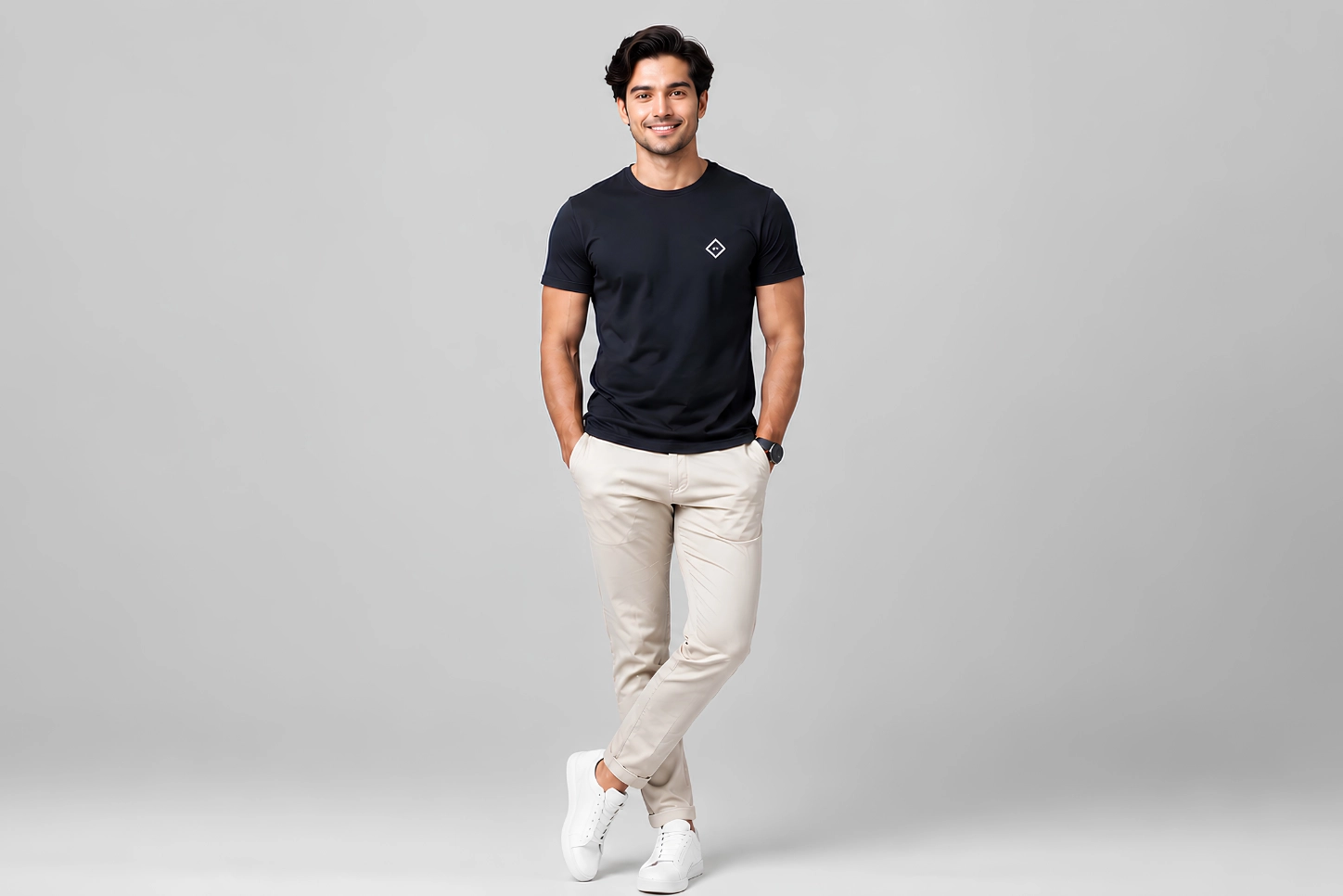 Man in black t-shirt and beige pants stands confidently, hands in pockets, against a gray background, smiling warmly.